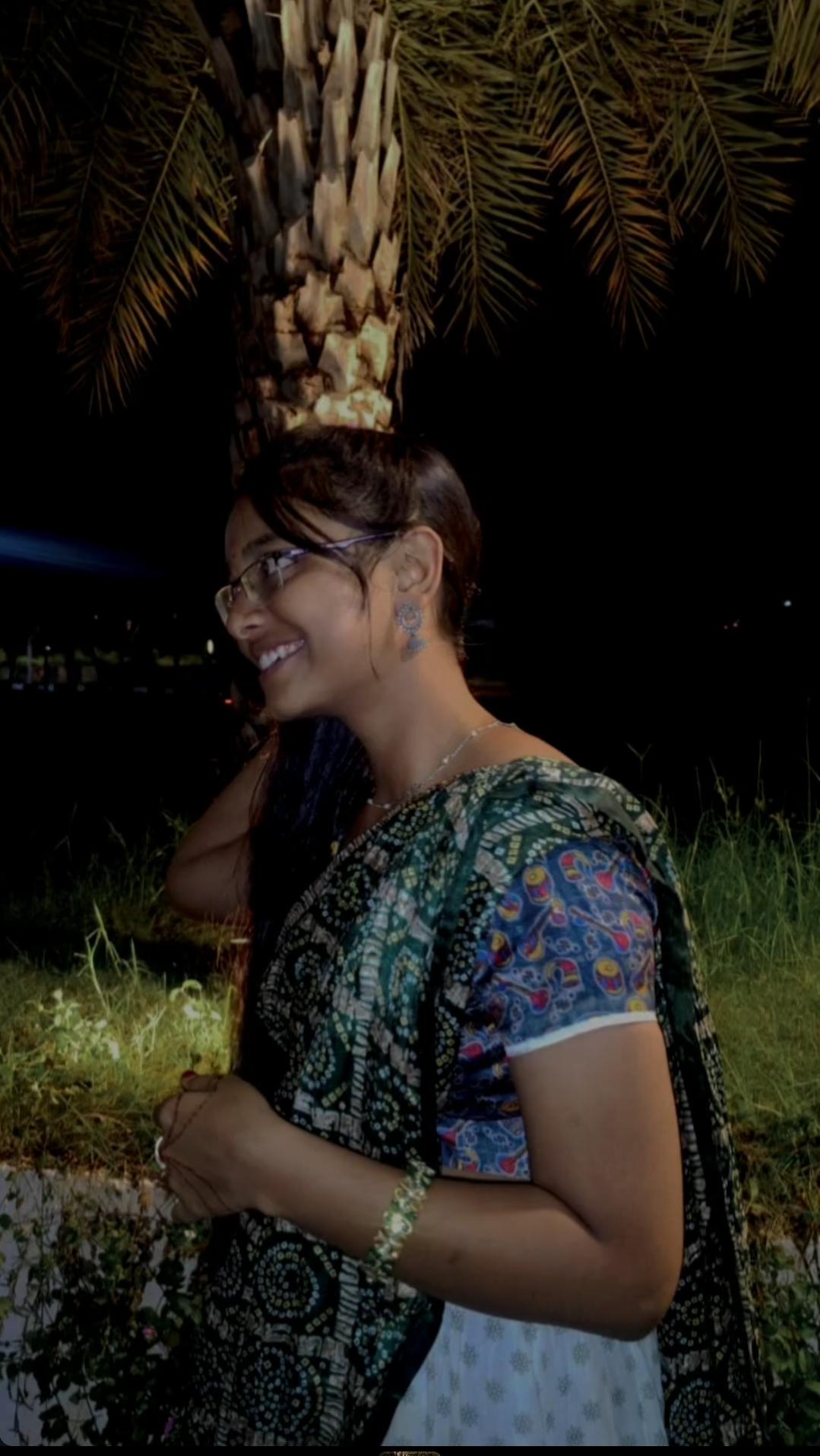 Jayani Bai smiling under a palm tree at night, in a saree